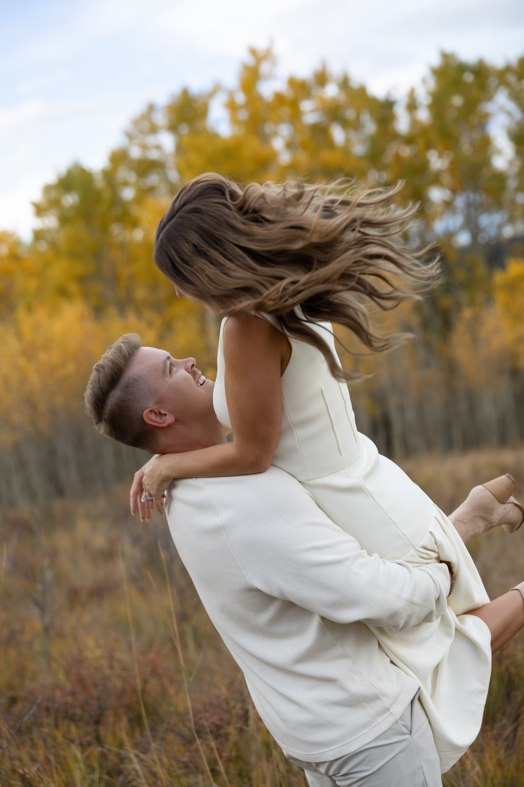 An engaged couple wearing all white where the man is lifting the woman up.